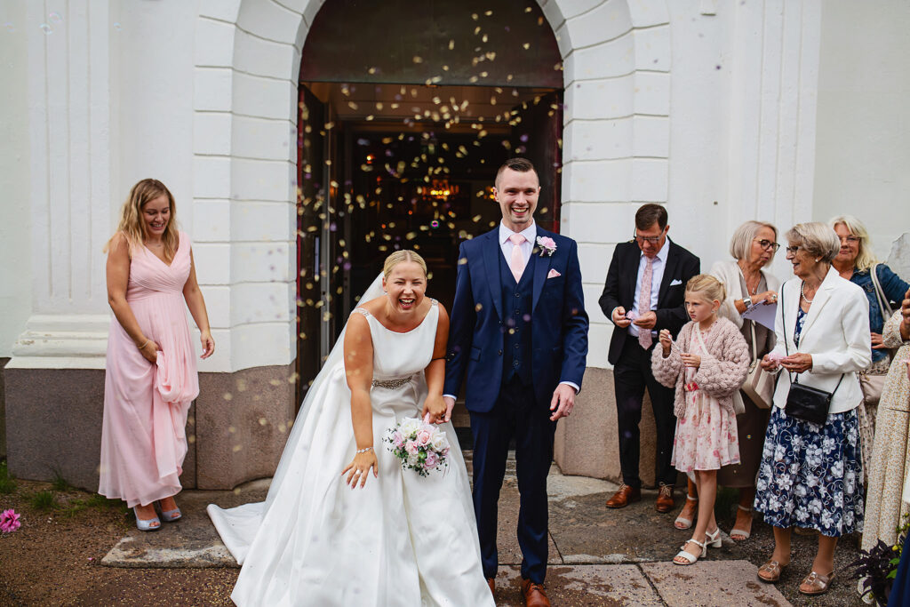 Bröllopsfotografering - brudparet har precis kommit ut ur kyrkan och gästerna kastar ris på dem. Bruden skrattar och strålar