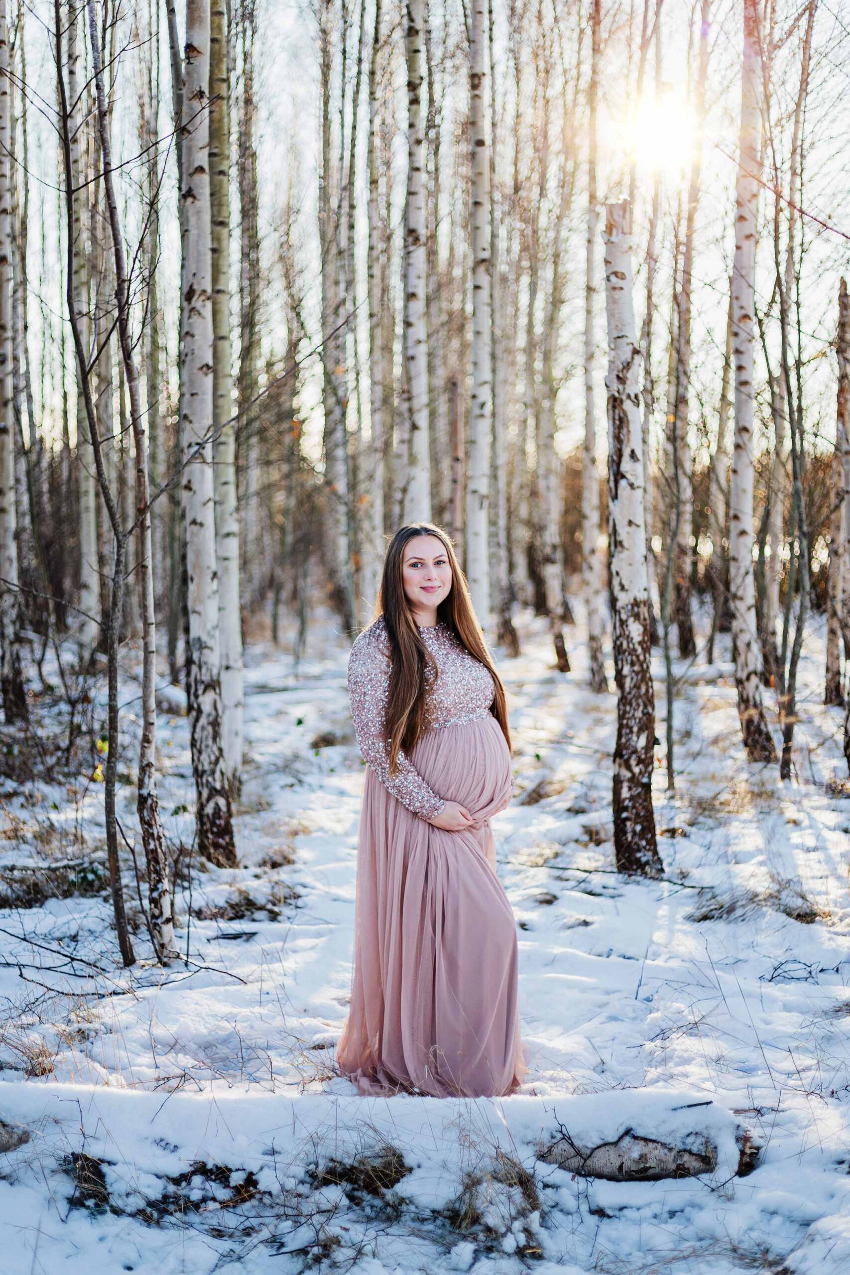 Gravid kvinna som står i skogen bland björkstammar. Marken är täckt av snö och solen tittar fram bakom björkarna. Hon har på sig en rosa klänning och håller båda händerna under magen och tittar in i kameran och ler.