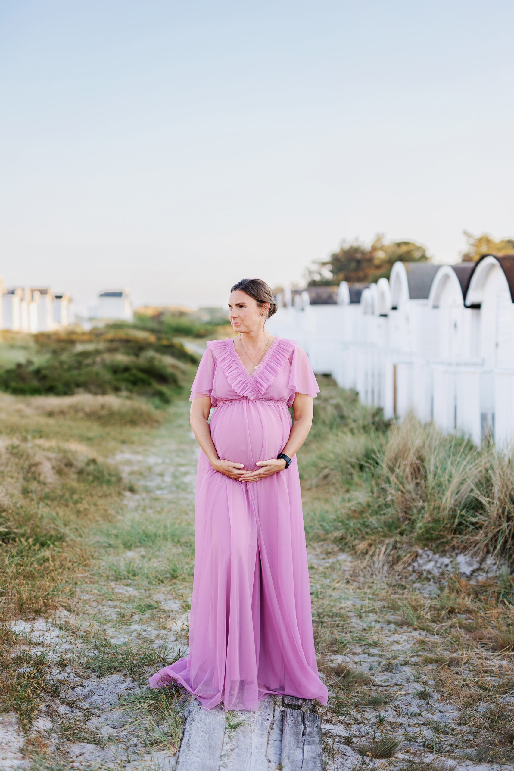 Gravid kvinna i fin klänning som håller under magen. Hon står en bit upp från stranden mellan strandhytter på en gång i trä.