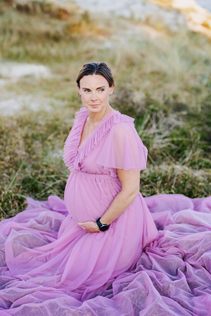 Gravid kvinna som sitter på knä i vass/gräs på stranden. Hon håller båda händerna under magen och tittar sidan om kameran.