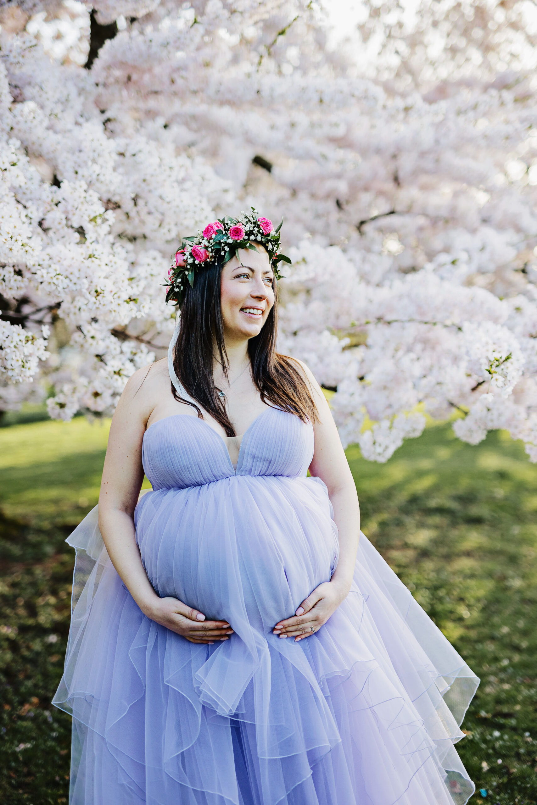 Gravid kvinna som står framför ett stort körsbärsträd i en fluffig klänning. Hon håller under magen, tittar åt sidan och har en blomsterkrans på huvudet.