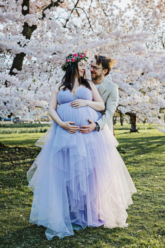 Ett par står framför ett stort körsbärsträd som blommar. Mannen står bakom den gravida kvinnan och håller om henne. De tittar på varandra och ler stort. Kvinnan har en stor klänning och blomsterkrans på sig.