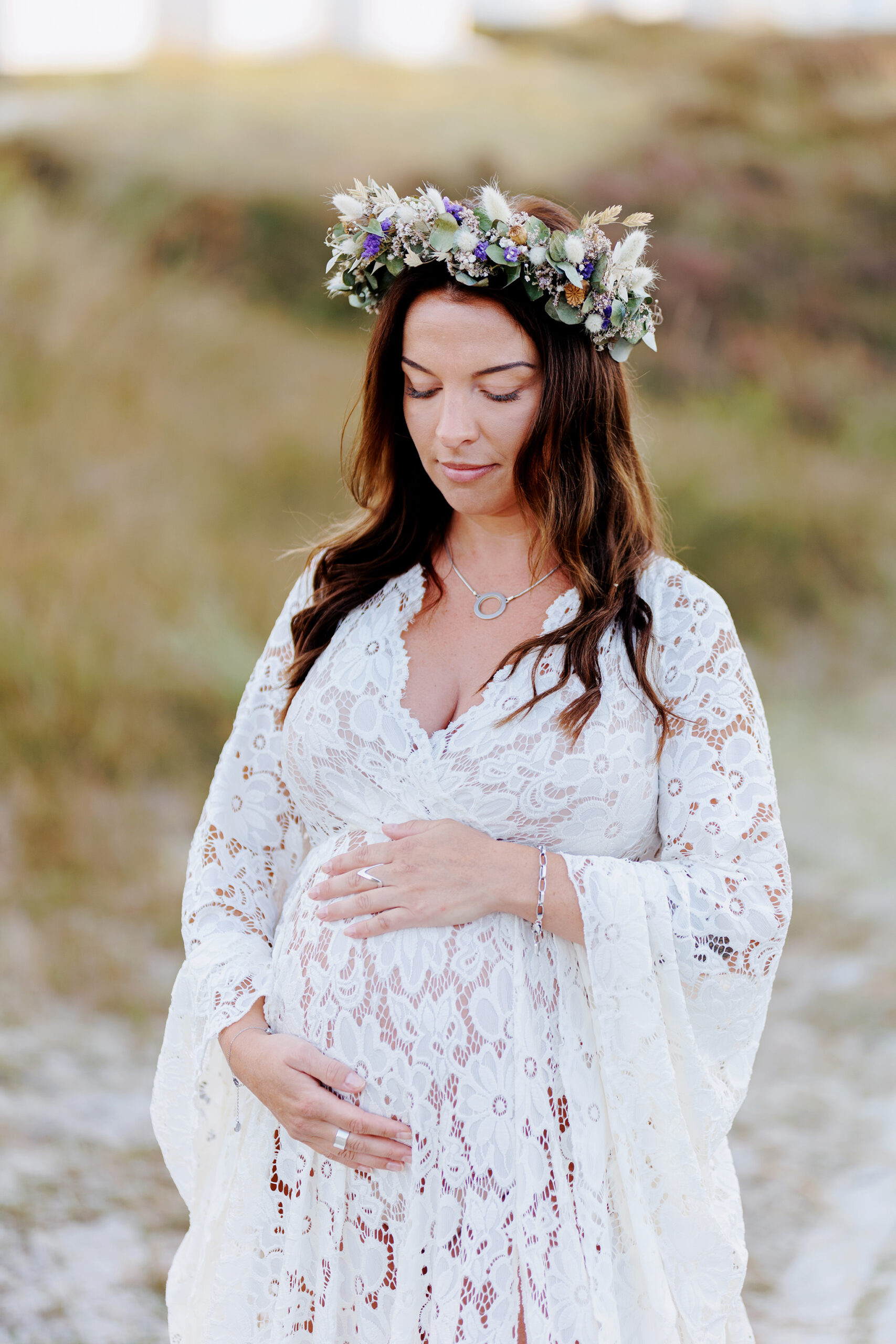 Halvkroppsbild på en gravid kvinna i spetsklänning som står och håller om sin mage. Hon har en blomsterkrans i håret.