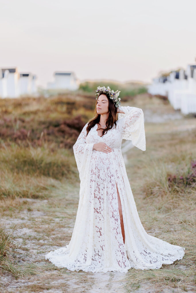 Gravid kvinna i spetsklänning som håller en handen på magen och andra handen i håret. Hon står en bit upp från stranden mellan strandhytter på en gång i trä och har en blomsterkrans på huvudet.