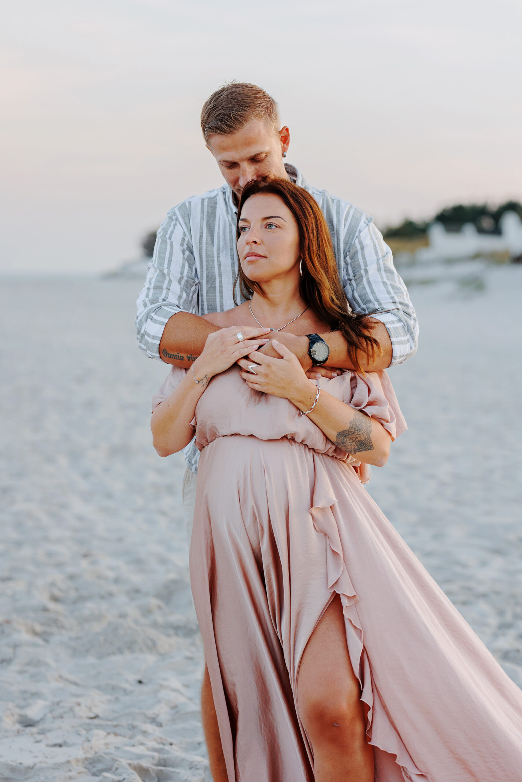 En man står bakom en gravid kvinna och håller om henne, samtidigt som han pussar henne i håret. Paret står på stranden och kvinnan tittar ut över havet.