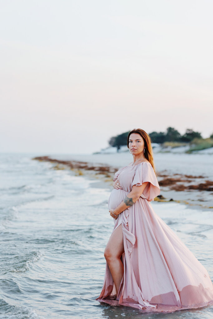 En gravid kvinna står i vattenbrynet på stranden med en klänning med långt släp som böljar i vattnet.