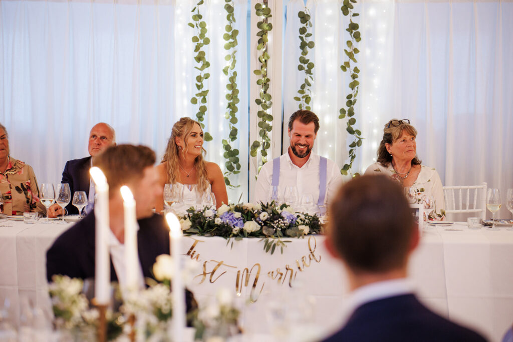 Bröllopsfotograf Skåne - brudpar som sitter vid ett fint dukat bord och skrattar, med gäster omkring sig.