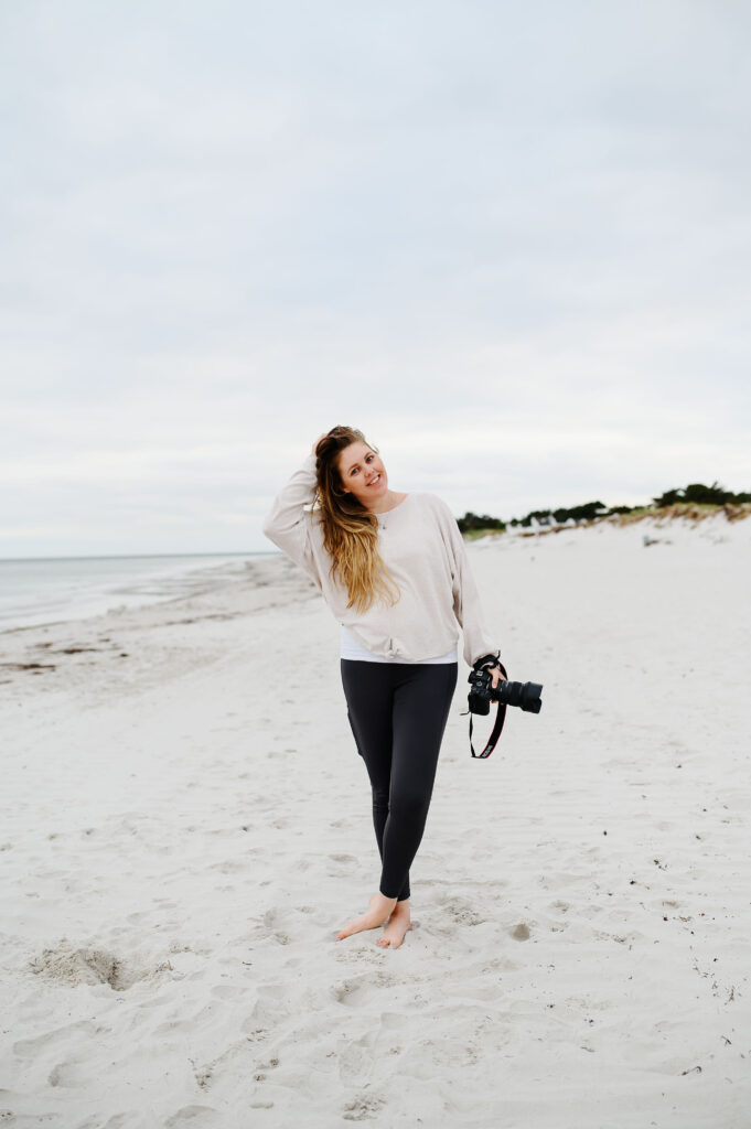 Bröllopsfotograf Skåne - en kvinna som står på en strand med en kamera i ena handen och den andra handen i håret och ler in i kameran.