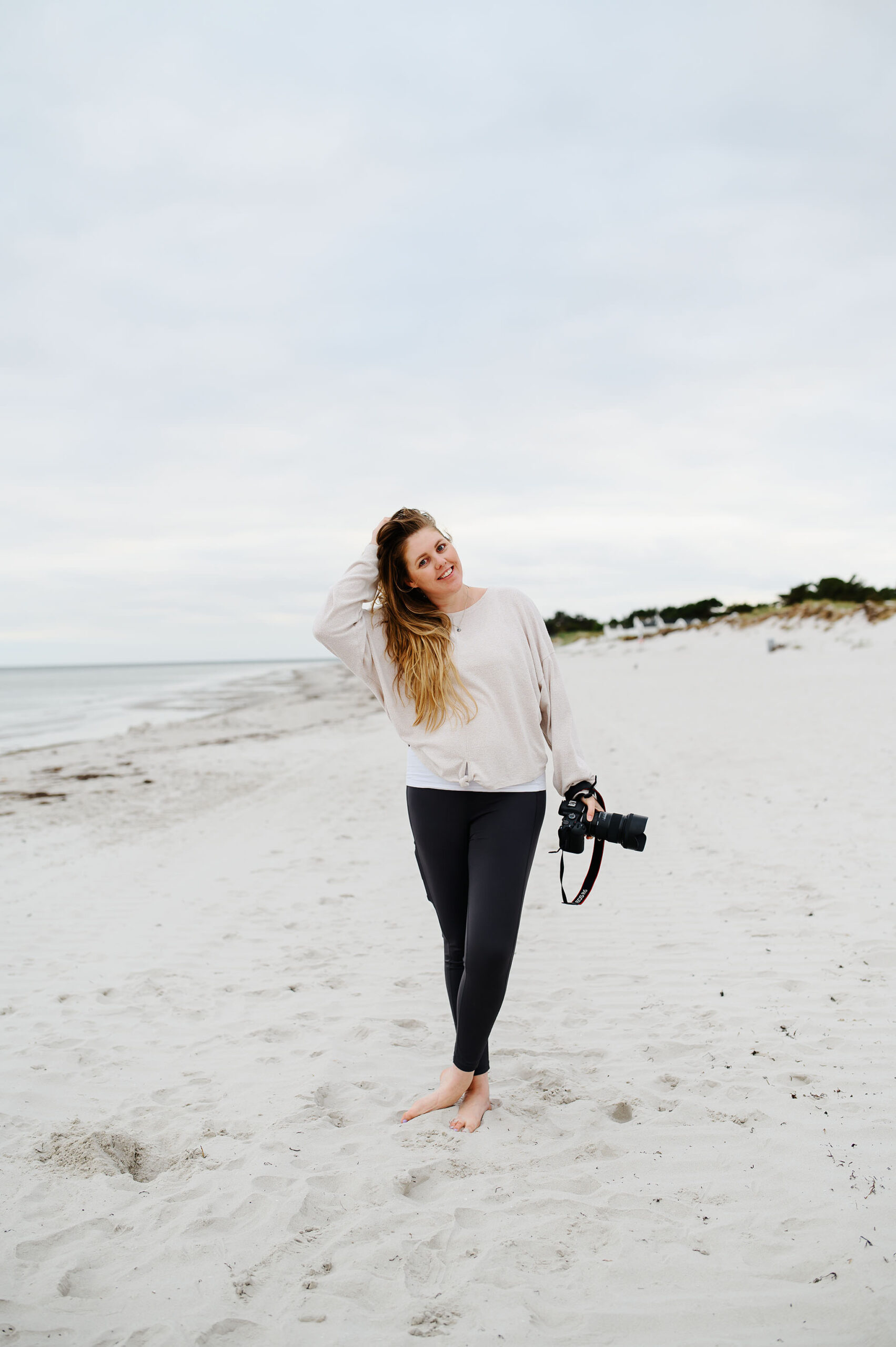 Bröllopsfotograf Skåne - en kvinna som står på en strand med en kamera i ena handen och den andra handen i håret och ler in i kameran.