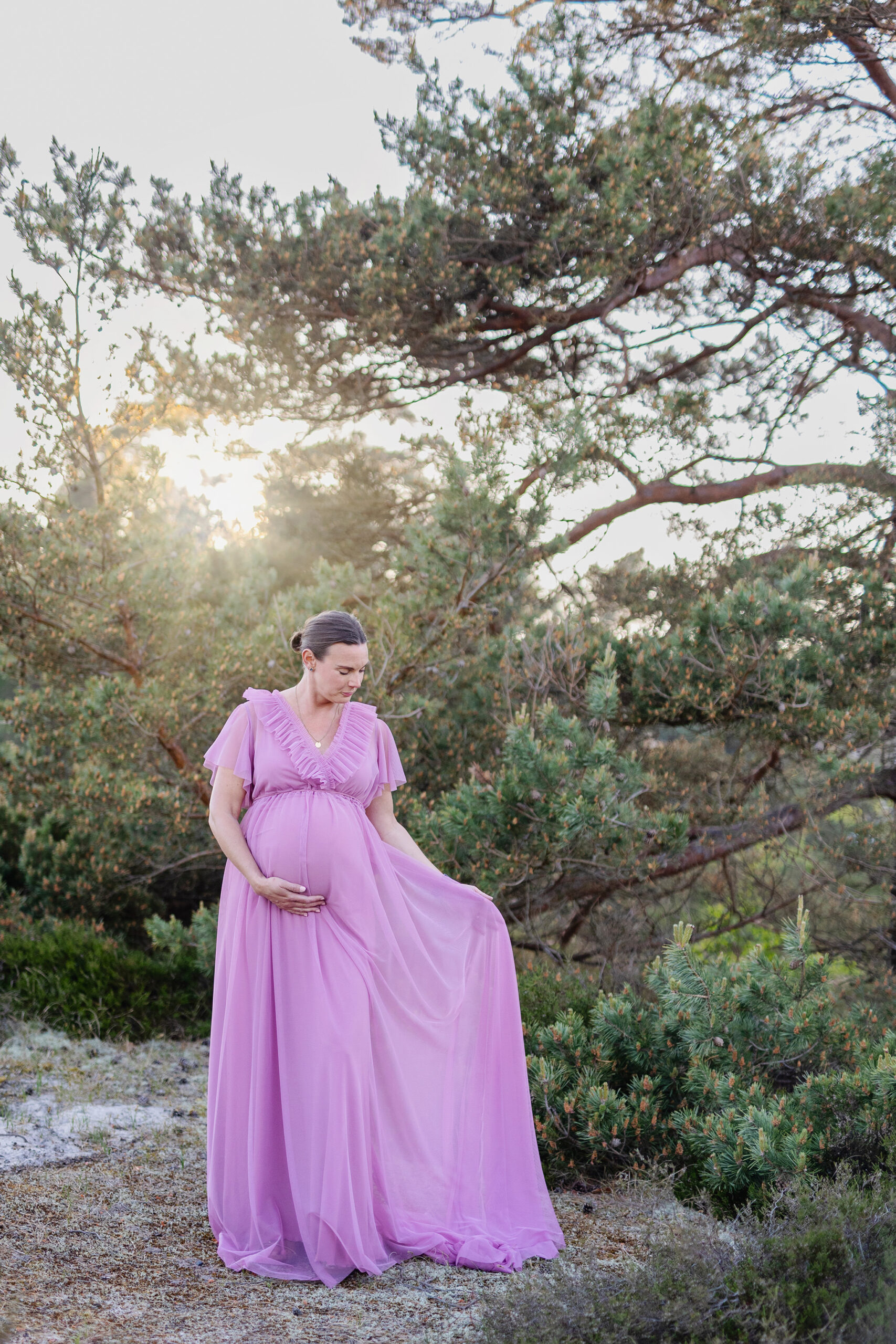 Gravidfotografering Malmö, Gravidfotografering Skåne Gravidfotografering Malmö - Gravid kvinna som står framför ett träd och håller ena handen i klänningen och andra under magen.