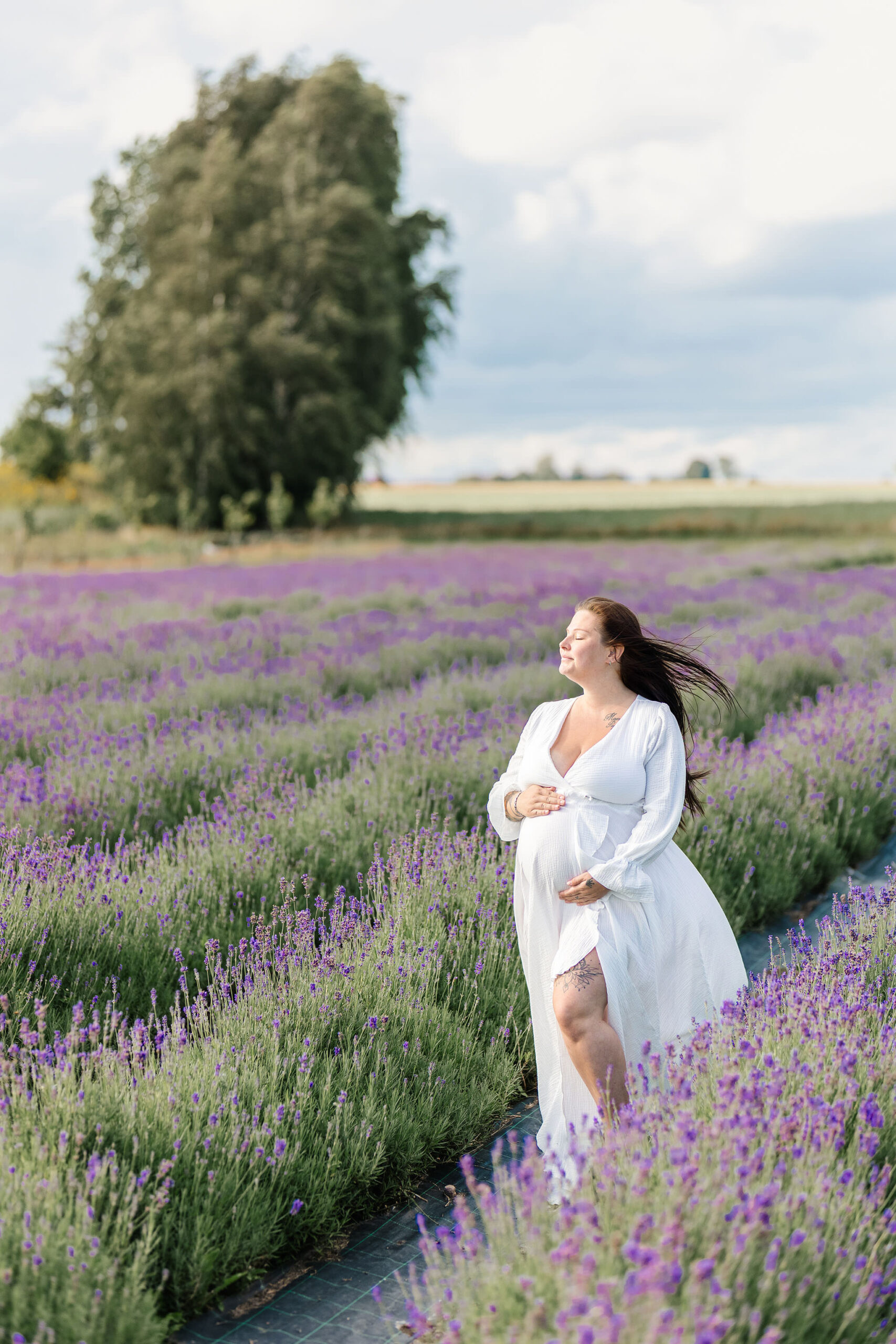 Gravidfotografering Malmö, Skåne - Gravid kvinna som står i en vit klänning och håller sin mage mitt i ett lavendelfält.