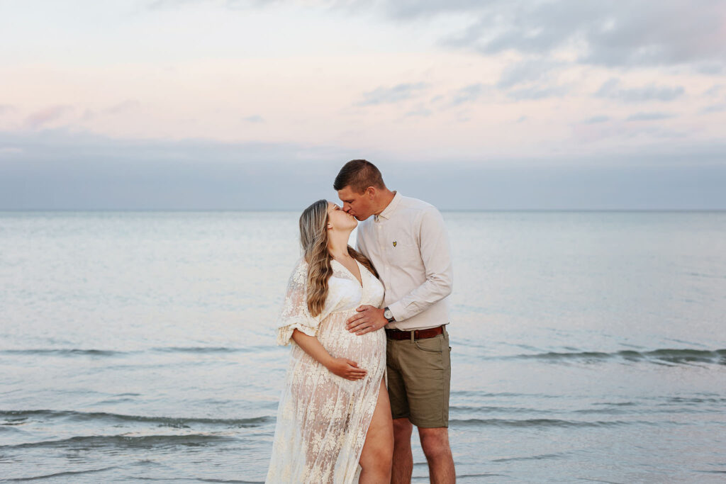 Gravidfotografering Malmö, Gravidfotografering Skåne - ett gravidpar som står på stranden och pussas. De håller båda kärleksfullt om magen.