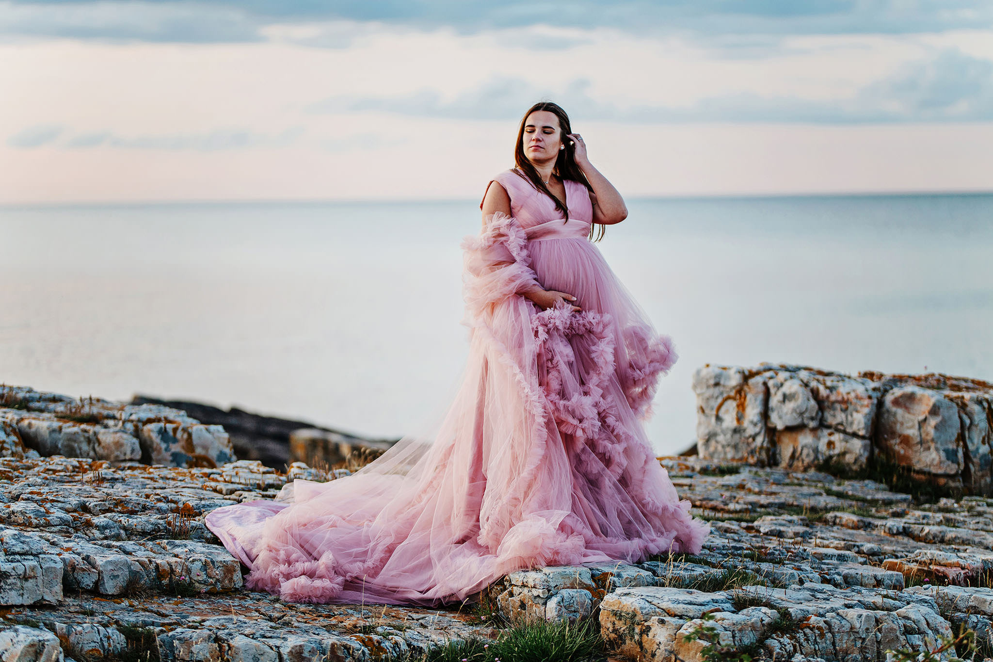 Gravidfotografering Malmö, Gravidfotografering Skåne - gravid kvinna som står på klippor ovanför vattnet i en pampig klänning och håller om sin mage.