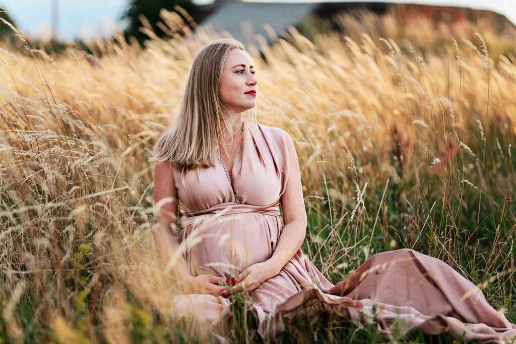 Gravidfotografering Malmö, Gravidfotografering Skåne - gravdi kvinna som sitter i högt gräs i en fin klänning och håller om magen.