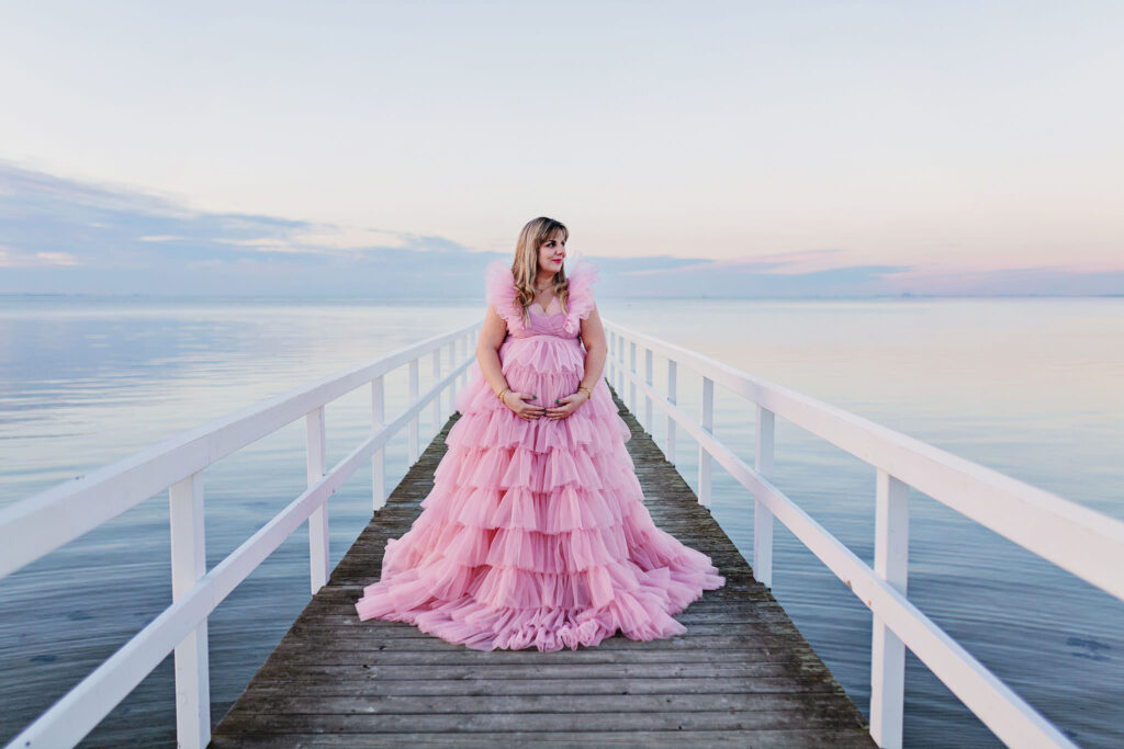 Gravidfotografering Malmö, Gravidfotografering Skåne En gravidkvinna som står på en bro med en stor, flyffig rosa klänning på sig. Hon håller om magen och tittar ut åt sidan.