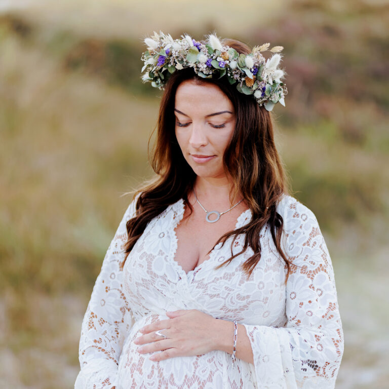 Gravidfotografering Malmö - Halvkroppsbild på en gravid kvinna i spetsklänning som står och håller om sin mage. Hon har en blomsterkrans i håret.