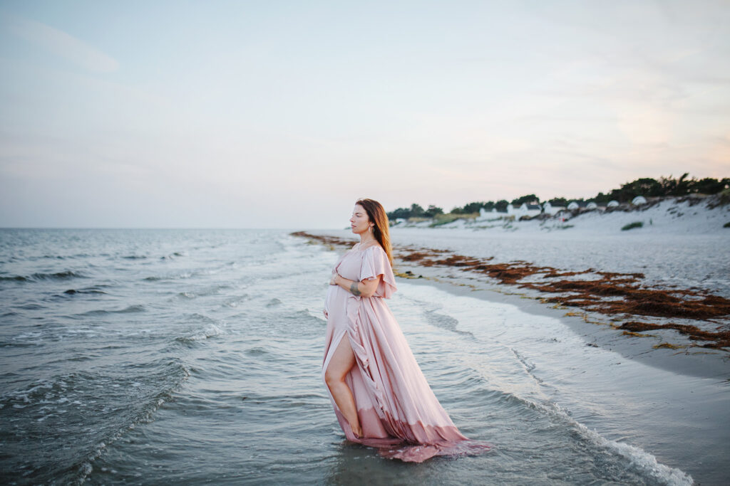 Gravidfotografering Malmö - En gravid kvinna står i vattenbrynet på stranden med en klänning med långt släp som böljar i vattnet.
