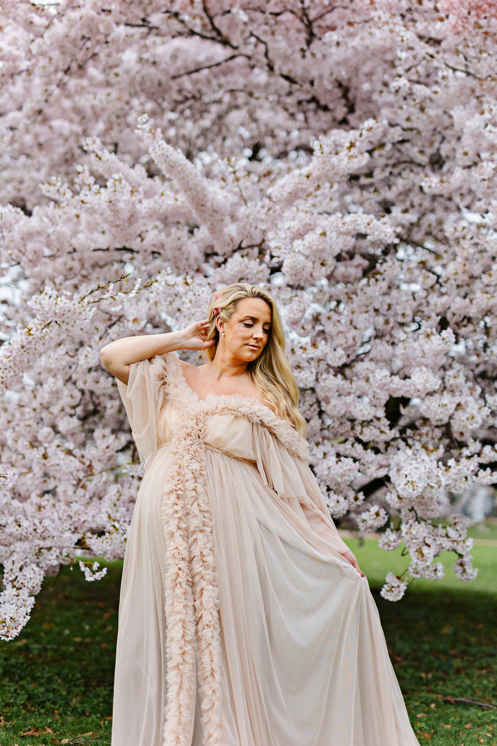 Gravidfotografering Malmö, Skåne - En gravid kvinna står framför ett stort körsbärsträd i en fluffig klänning. Hon håller ena handen i håret och andra handen håller i klänningen.